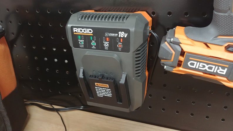 May include: A black and orange RIDGID 18V lithium-ion battery charger with a green light indicating charging. The charger is mounted on a black pegboard.