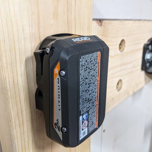 May include: A black and orange 18V 2Ah lithium-ion battery for a power tool. The battery is mounted on a wooden wall with a metal bracket. The battery has the word "RIDGID" printed on it.
