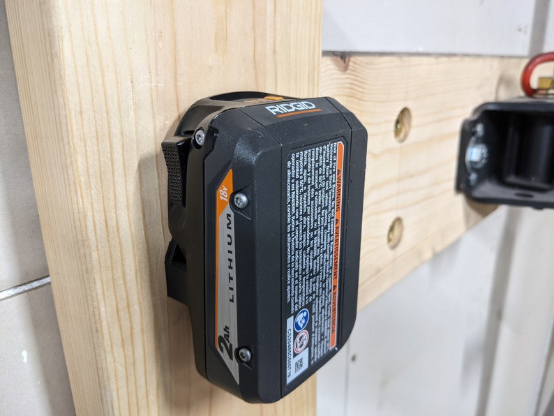 May include: A black and orange 18V 2Ah lithium-ion battery for a power tool. The battery is mounted on a wooden wall with a metal bracket. The battery has the word "RIDGID" printed on it.
