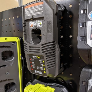 May include: A black and yellow Ryobi battery charger with a black power cord. The charger has a label that says "IntelliPort" and has a red and yellow light indicator. The charger is mounted on a black pegboard.