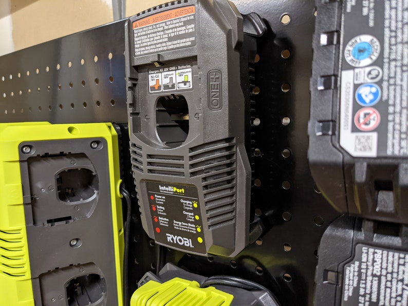 May include: A black and yellow Ryobi battery charger with a black power cord. The charger has a label that says "IntelliPort" and has a red and yellow light indicator. The charger is mounted on a black pegboard.