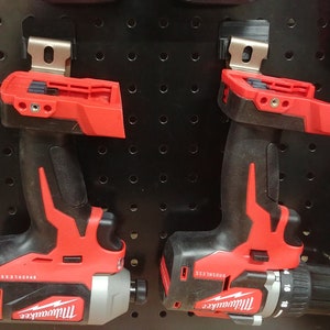 May include: Two Milwaukee power tools, a red and black impact driver and a red and black drill, are hanging on a black pegboard. Both tools have a silver metal bit holder.