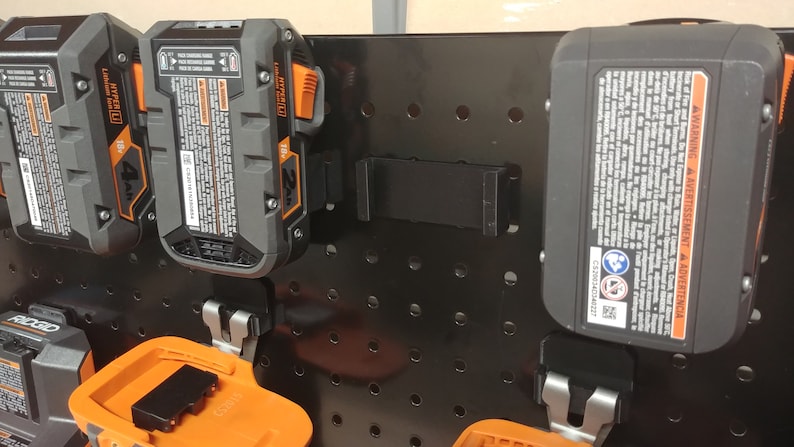 May include: A black pegboard with multiple orange and black battery packs hanging on it. The battery packs are labeled "2Ah" and "4Ah".
