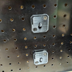 May include: Close-up of a black pegboard with evenly spaced holes. Two gray, 3D-printed pegboard mounting brackets are attached with screws. The brackets have a rectangular shape with a central peg for hanging items.