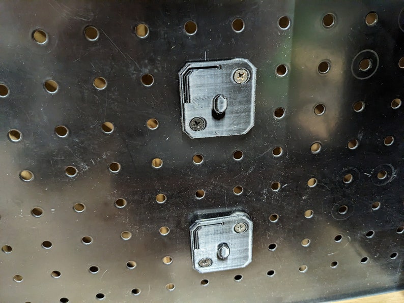 May include: Close-up of a black pegboard with evenly spaced holes. Two gray, 3D-printed pegboard mounting brackets are attached with screws. The brackets have a rectangular shape with a central peg for hanging items.