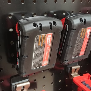 May include: Two black Milwaukee 12.0 Volt Lithium-Ion battery packs with red accents are hanging on a black pegboard.