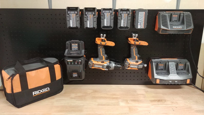Op de afbeelding: Een verzameling elektrisch gereedschap en accessoires, waaronder boormachines, opladers en batterijen, weergegeven op een zwart pegboard. De gereedschappen zijn voornamelijk grijs en oranje, met de merknaam "RIDGID" zichtbaar. Er is ook een zwart-oranje gereedschapstas aanwezig.