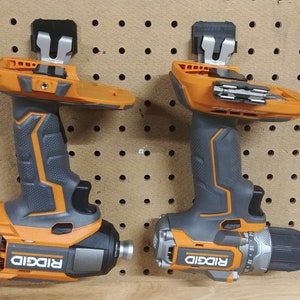 Op de afbeelding: Twee oranje en grijze elektrische boormachines met de merknaam "RIDGID" erop. Ze zijn gemonteerd op een pegboard. E&eacute;n boormachine heeft een boor, de andere een schroefbit.