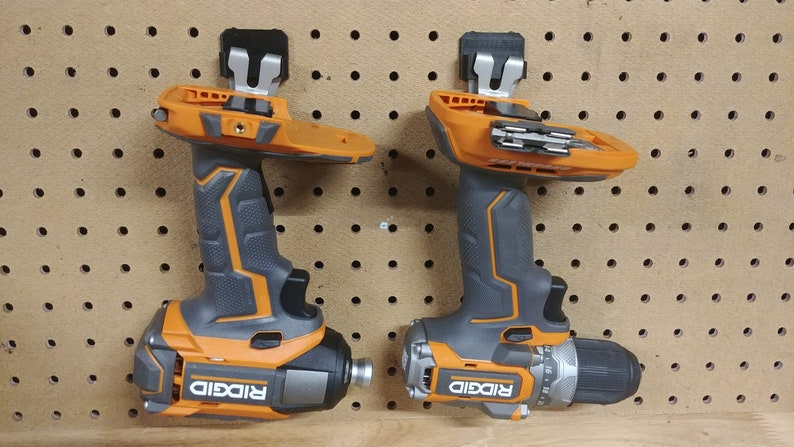 Op de afbeelding: Twee oranje en grijze elektrische boormachines met de merknaam "RIDGID" erop. Ze zijn gemonteerd op een pegboard. E&eacute;n boormachine heeft een boor, de andere een schroefbit.