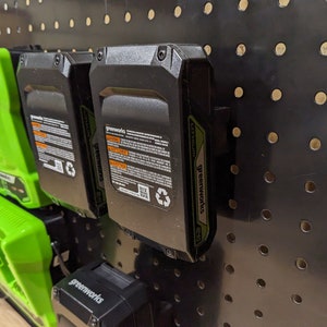 Może przedstawiać: Trzy czarne i zielone baterie litowo-jonowe do narzędzi elektrycznych są zamontowane na czarnej tablicy perforowanej. Baterie są oznaczone "Greenworks".