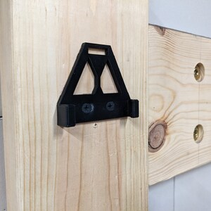 May include: A black plastic wall mount with two screw holes. The mount is shaped like a triangle with a rounded top and a flat bottom. It is attached to a wooden surface.