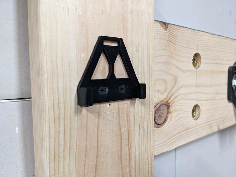 May include: A black plastic wall mount with two screw holes. The mount is shaped like a triangle with a rounded top and a flat bottom. It is attached to a wooden surface.