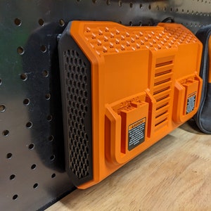 May include: An orange and black power tool battery charger mounted on a black pegboard. The charger has a honeycomb vent design and two battery slots. A black and orange bag is visible in the background.