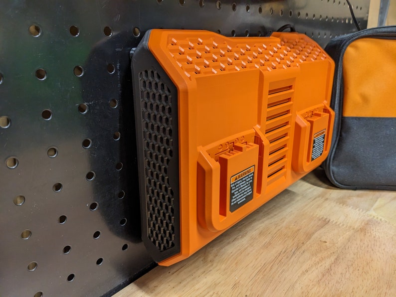 May include: An orange and black power tool battery charger mounted on a black pegboard. The charger has a honeycomb vent design and two battery slots. A black and orange bag is visible in the background.