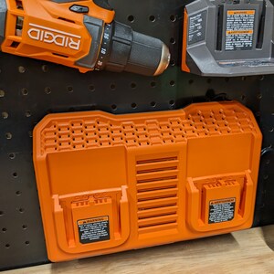 May include: Orange and black power tool accessories, including a drill, battery charger, and dual-bay battery charger. The drill is labeled "RIDGID". The battery charger has a warning label. The items are mounted on a black pegboard.
