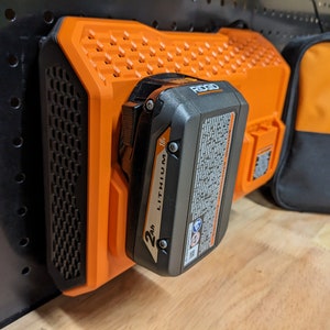 May include: An orange and black Ridgid 18V Lithium 2Ah battery pack attached to an orange charger. The charger is mounted on a black pegboard. A black and orange Ridgid tool bag is in the background.