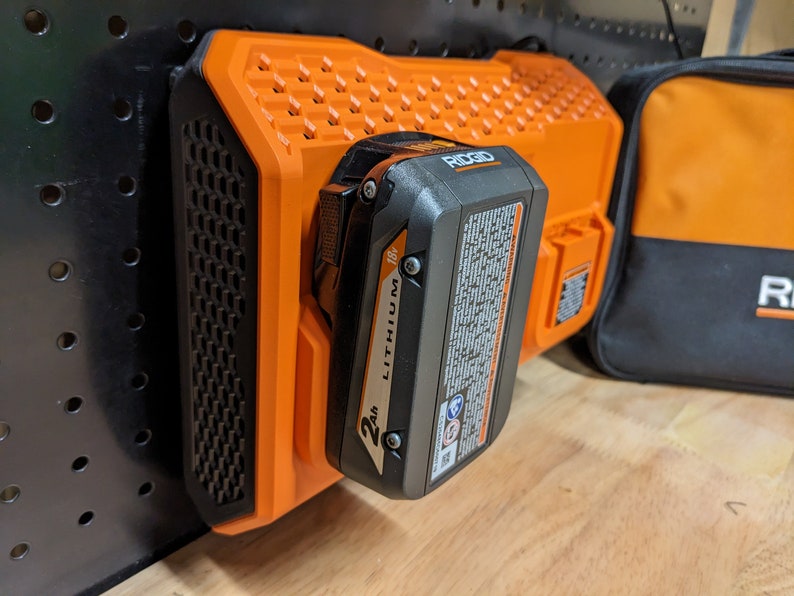 May include: An orange and black Ridgid 18V Lithium 2Ah battery pack attached to an orange charger. The charger is mounted on a black pegboard. A black and orange Ridgid tool bag is in the background.