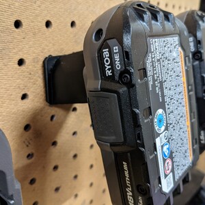 May include: Two black Ryobi ONE+ 18V lithium-ion batteries mounted on a pegboard wall. The batteries have a black and blue label with safety information and warnings.