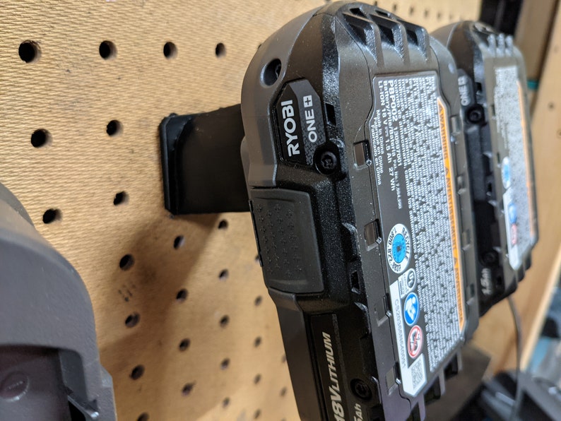 May include: Two black Ryobi ONE+ 18V lithium-ion batteries mounted on a pegboard wall. The batteries have a black and blue label with safety information and warnings.