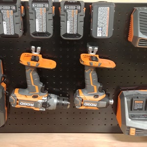Op de afbeelding: Een verzameling elektrisch gereedschap en opladers, waaronder boormachines en batterijpakketten, gemonteerd op een zwart pegboard. De gereedschappen zijn voornamelijk grijs en oranje, met de merknaam "RIDGID".
