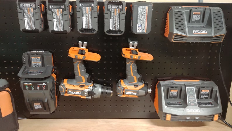 Op de afbeelding: Een verzameling elektrisch gereedschap en opladers, waaronder boormachines en batterijpakketten, gemonteerd op een zwart pegboard. De gereedschappen zijn voornamelijk grijs en oranje, met de merknaam "RIDGID".