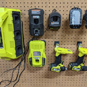 May include: A wall-mounted organizer with multiple Ryobi power tool chargers and two Ryobi drill drivers. The chargers are green and black, and the drill drivers are yellow and black.