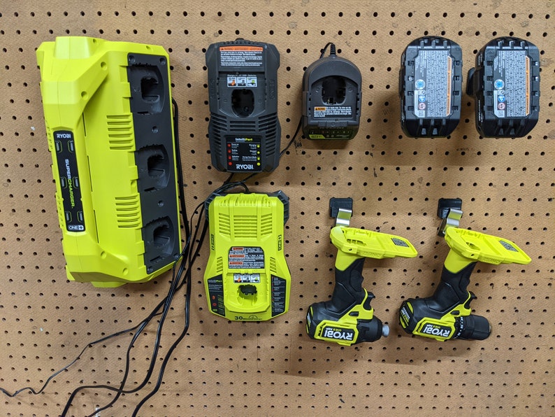 May include: A wall-mounted organizer with multiple Ryobi power tool chargers and two Ryobi drill drivers. The chargers are green and black, and the drill drivers are yellow and black.