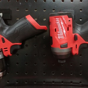 May include: Two Milwaukee FUEL power tools, a red and black drill and a red and black impact driver, are hanging on a black pegboard.