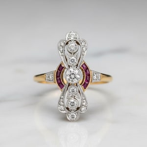 Peut inclure: Une bague en or avec une pièce centrale verticale et allongée en métal blanc. Le design comprend un diamant rond central, des diamants plus petits en accent et deux rubis rouge foncé incurvés placés de chaque côté de la pierre centrale.