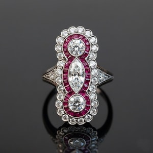 May include: An Art Deco-style ring with a central marquise-cut diamond, flanked by two round diamonds and surrounded by a halo of small rubies and diamonds. The ring has a silver-colored band and intricate detailing.