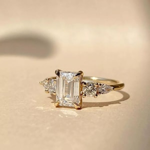 Op de afbeelding: Een gouden ring met een grote, rechthoekig geslepen diamant in het midden, geflankeerd door kleinere, peervormige diamanten aan weerszijden. De ring heeft een delicate band en een klassiek, elegant ontwerp.