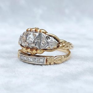 Puede incluir: Dos anillos de oro antiguos. El anillo superior presenta un diamante central y detalles ornamentados en plata y oro. El anillo inferior tiene una banda de pequeños diamantes y detalles florales. Ambos anillos están sobre un fondo blanco suave.