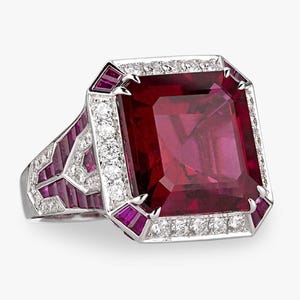 May include: A striking ring featuring a large, rectangular, deep red gemstone. The ring is set in white metal and surrounded by a halo of small diamonds and smaller, rectangular red gemstones. The design is geometric.