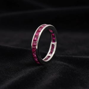 May include: A silver-toned eternity band ring featuring a continuous channel setting of square-cut, vibrant magenta-red gemstones. The metal band has a polished, reflective finish that contrasts with the deep red color of the inset stones.
