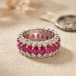 May include: A silver-toned eternity band featuring a central row of oval-cut vibrant pink stones flanked by two rows of smaller, round-cut clear stones. The ring has a metallic setting that holds the stones in a continuous circular pattern.