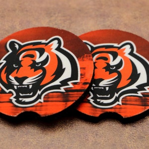 May include: Two round orange and black coasters with a tiger head graphic. The tiger has a white mouth and white stripes. The background is a blurred image of a stadium.