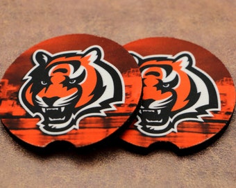 Puede incluir: Dos posavasos redondos de color naranja y negro con un gráfico de cabeza de tigre. El tigre tiene la boca blanca y rayas blancas. El fondo es una imagen borrosa de un estadio.