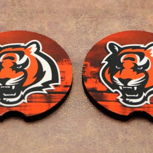 May include: Two round orange, black, and white coasters with a tiger head graphic. The tiger has a black nose, white teeth, and black eyes. The background is a blurry image of a city skyline.