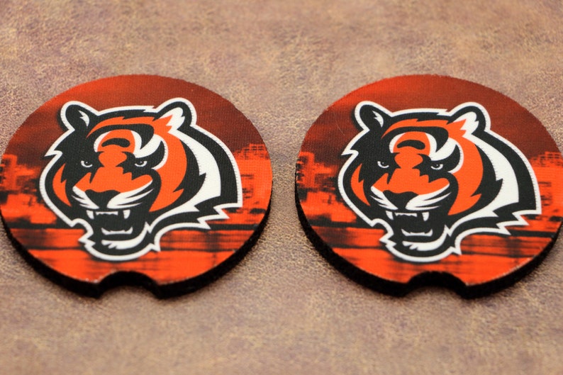 May include: Two round orange, black, and white coasters with a tiger head graphic. The tiger has a black nose, white teeth, and black eyes. The background is a blurry image of a city skyline.