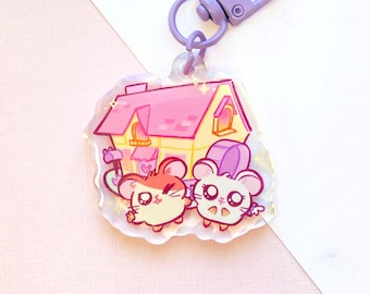 Könnte beinhalten: Ein lila-weißer Schlüsselanhänger mit einem Cartoon-Haus mit zwei Hamstern davor. Das Haus hat ein rosafarbenes Dach und gelbe Wände. Der Schlüsselanhänger ist aus klarem Acryl.