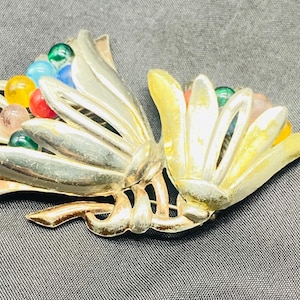 May include: A silver and gold tone brooch with a cluster of colorful glass stones in the center. The brooch is shaped like two stylized flowers.