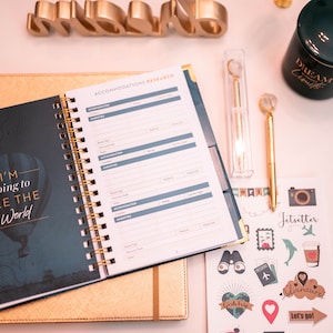 May include: A gold-colored planner with a spiral binding, open to a page with "I'm going to see the world" text and accommodation research sections. A gold pen, stickers, and a teal mug are also visible.