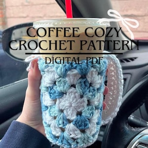 May include: A crocheted coffee cozy in a granny square pattern, featuring shades of blue and white. The cozy has a handle and is wrapped around a clear plastic cup with a red straw. Text on the image reads "COFFEE COZY CROCHET PATTERN DIGITAL PDF."