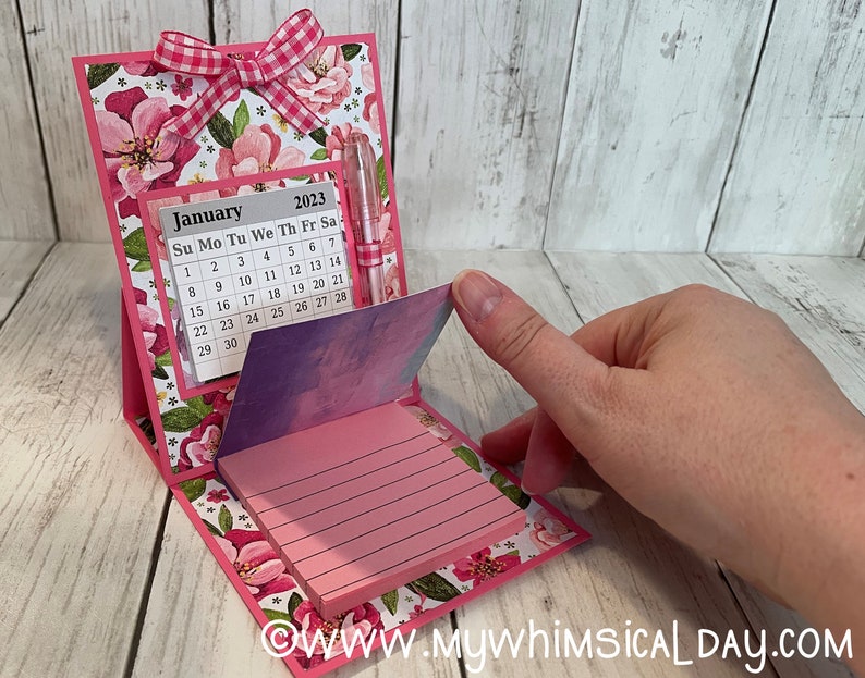 May include: A pink floral desktop calendar with a notepad and pen. The calendar is for January 2023 and has a pink gingham bow on top.