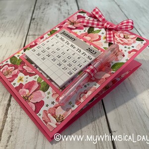 May include: A pink floral patterned card with a calendar for January 2023 and a pink pen with a white and pink gingham ribbon. The card has a white and pink gingham bow.