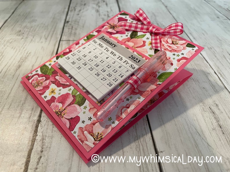 May include: A pink floral patterned card with a calendar for January 2023 and a pink pen with a white and pink gingham ribbon. The card has a white and pink gingham bow.