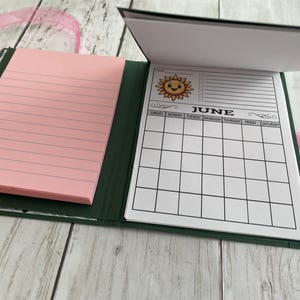 Puede incluir: Un bloc de notas verde y rosa con un calendario para el mes de junio. El calendario presenta un gr&aacute;fico de sol sonriente y la palabra "junio" en una fuente decorativa. El bloc de notas tiene p&aacute;ginas rayadas para escribir notas.