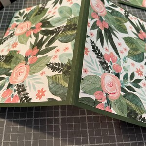 Puede incluir: Una tarjeta doblada con un borde verde y un fondo blanco con un patr&oacute;n floral con flores rosas y hojas verdes.