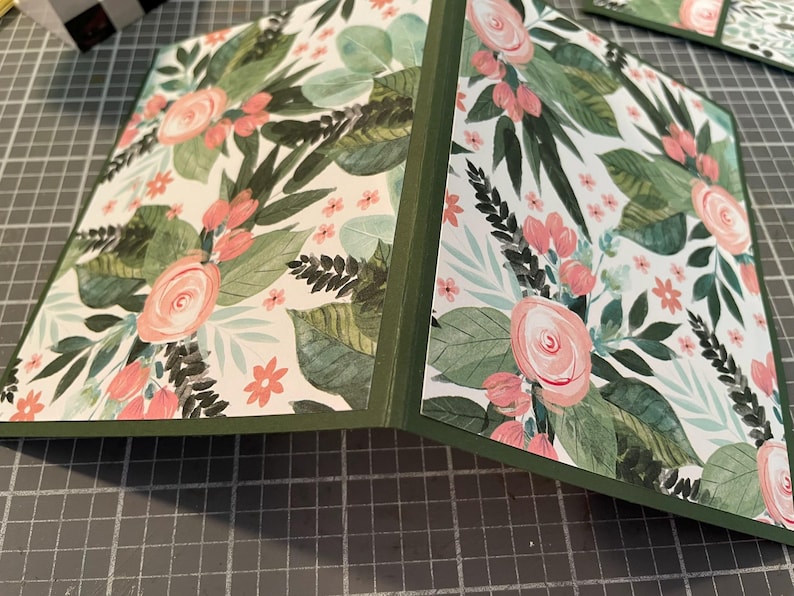 Puede incluir: Una tarjeta doblada con un borde verde y un fondo blanco con un patr&oacute;n floral con flores rosas y hojas verdes.