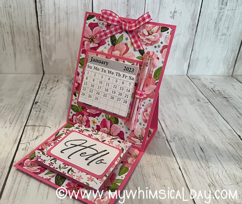May include: A pink floral desktop calendar with a notepad and pen. The calendar is for January 2023 and has a pink gingham bow on top. The notepad says "Hello".